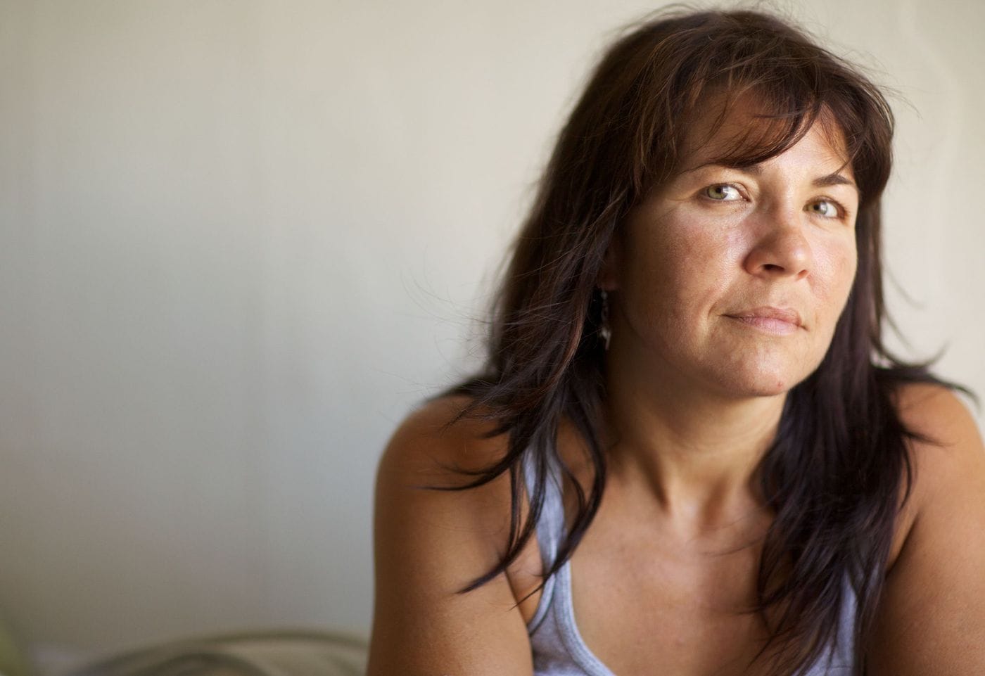 A close-up portrait of a woman with dark brown hair and bangs looking directly at the camera with a gentle expression against a plain, light background.