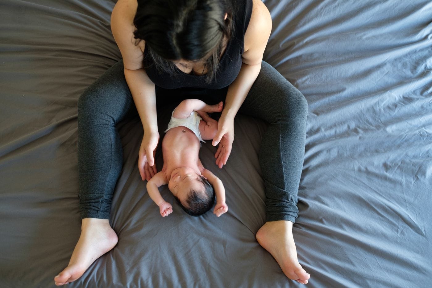 A top-down view shows a woman sitting on a gray bed sheet, gently tending to her newborn baby, who lies in a diaper between her legs with arms raised.