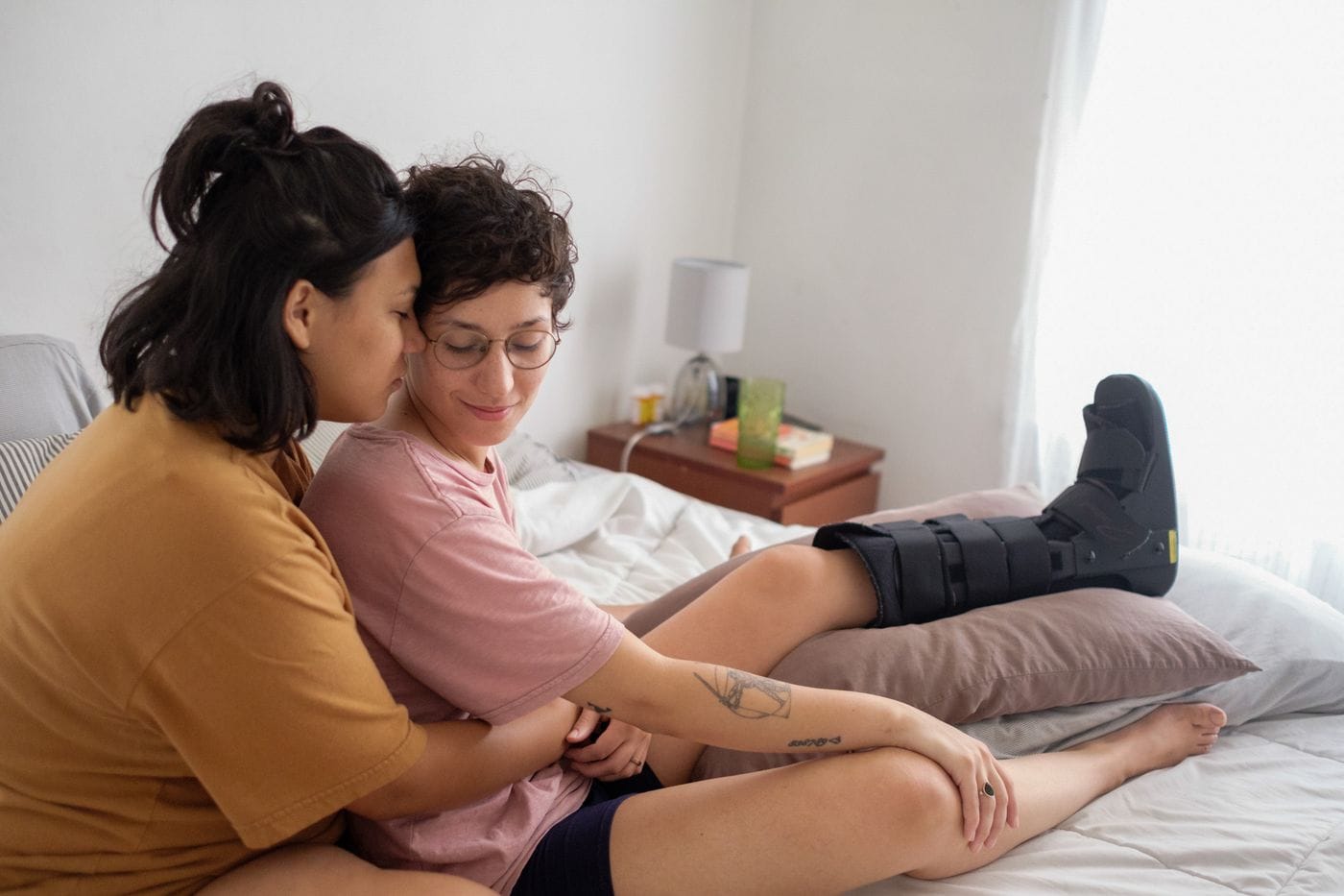 Two people sit closely together on a bed, with one resting their head against the other’s shoulder while the other, wearing a medical boot on their raised leg, looks down with a gentle smile.