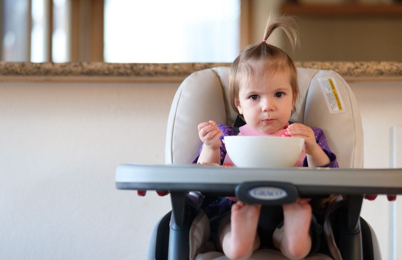 A toddler with a small ponytail in a high chair gazes forward while holding food in both hands in front of a white bowl.