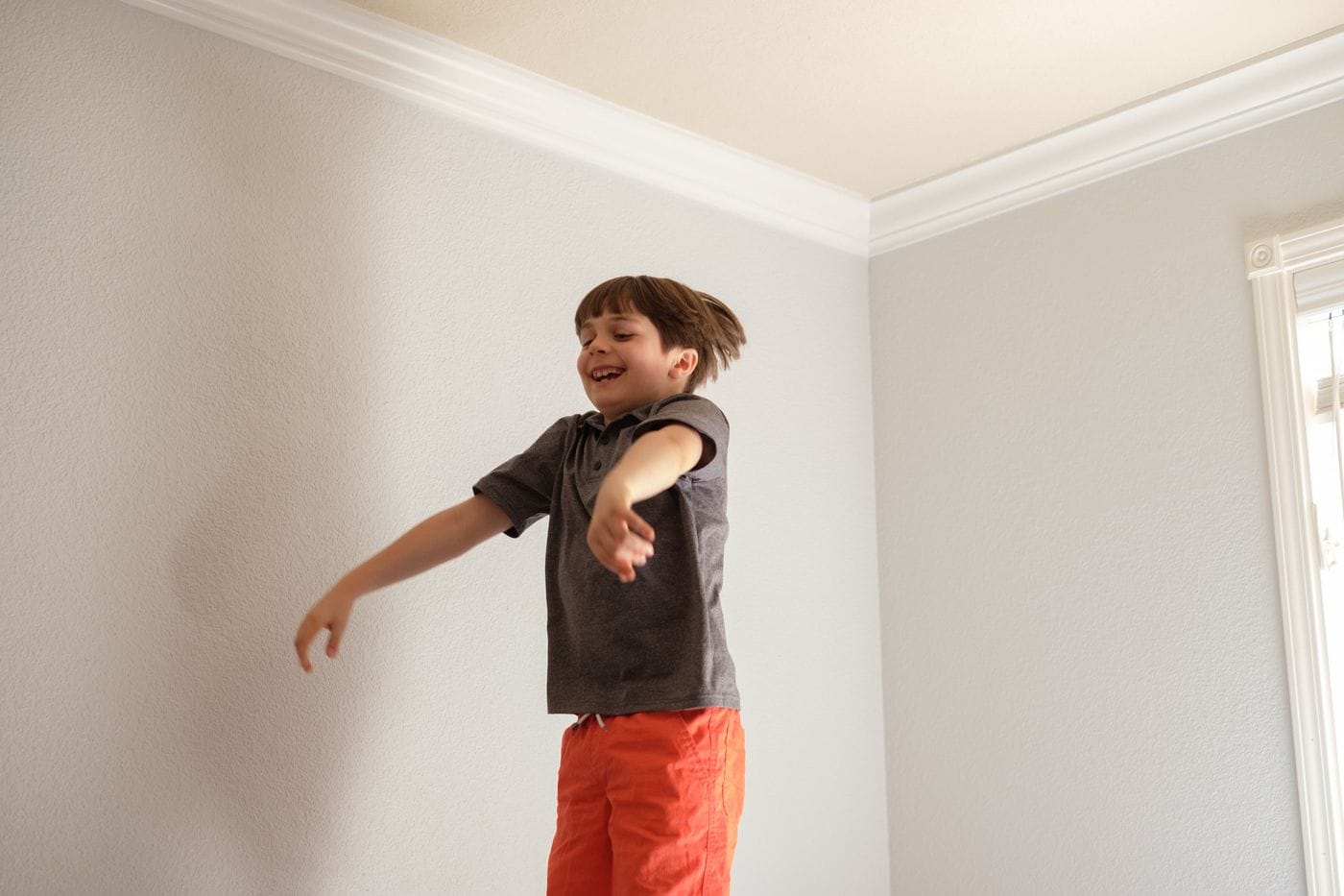 A young boy with brown hair, wearing a gray polo shirt and orange shorts, jumps in mid-air in a room with light gray walls and white trim.