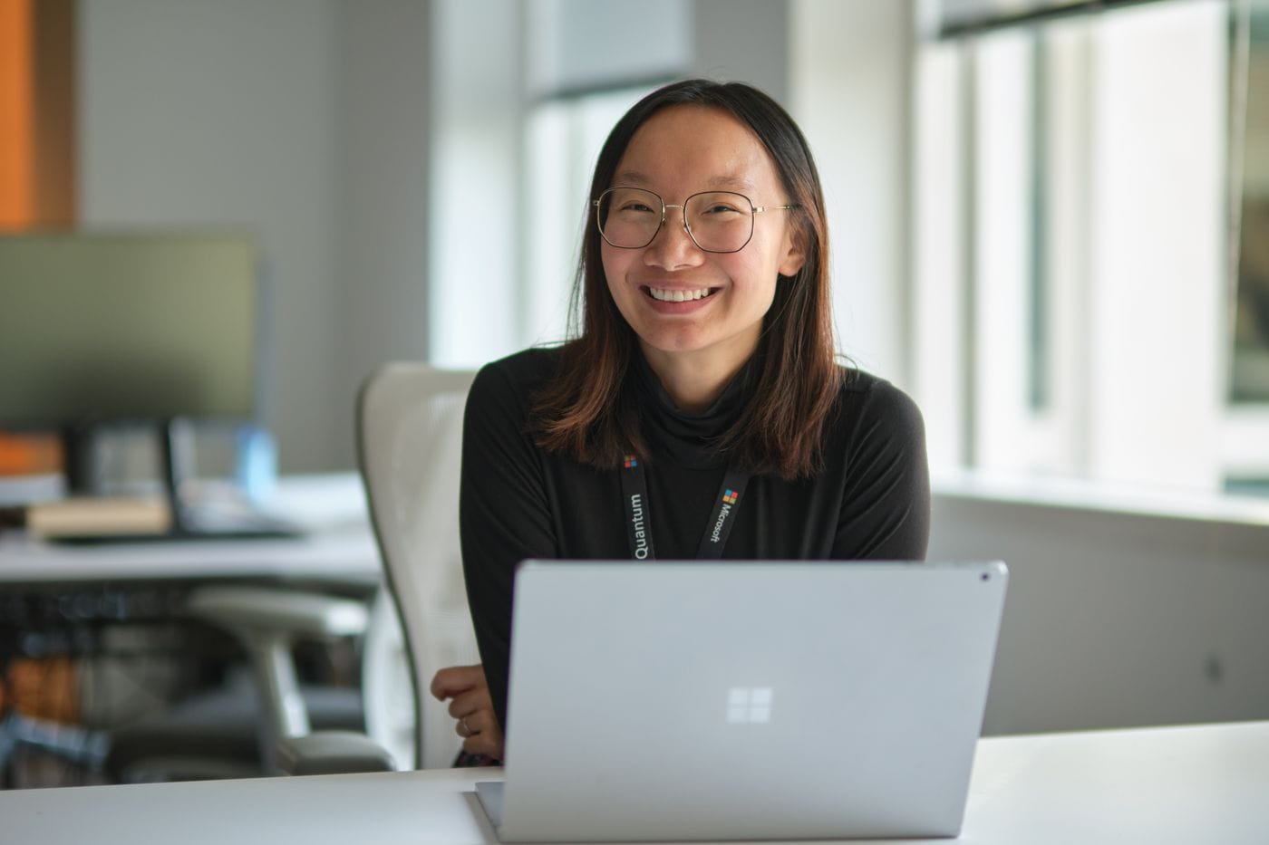 A smiling woman wearing glasses and a black turtleneck sits at a desk with a Microsoft laptop and a Microsoft Quantum lanyard in a modern office.