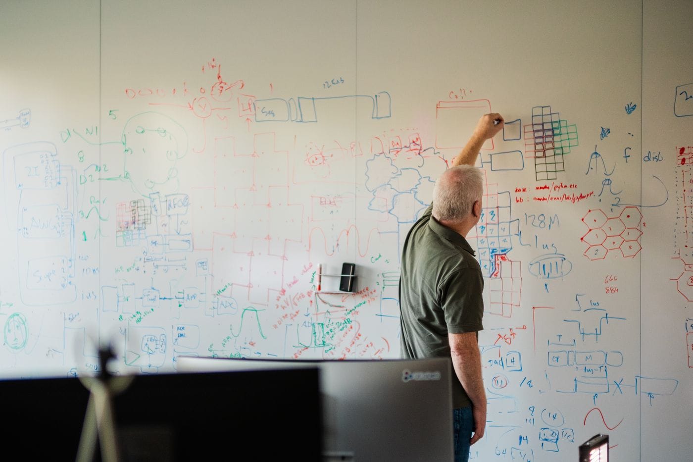 An older man with gray hair in a green polo shirt stands facing a large whiteboard, drawing diagrams and writing technical notes in multiple colors.
