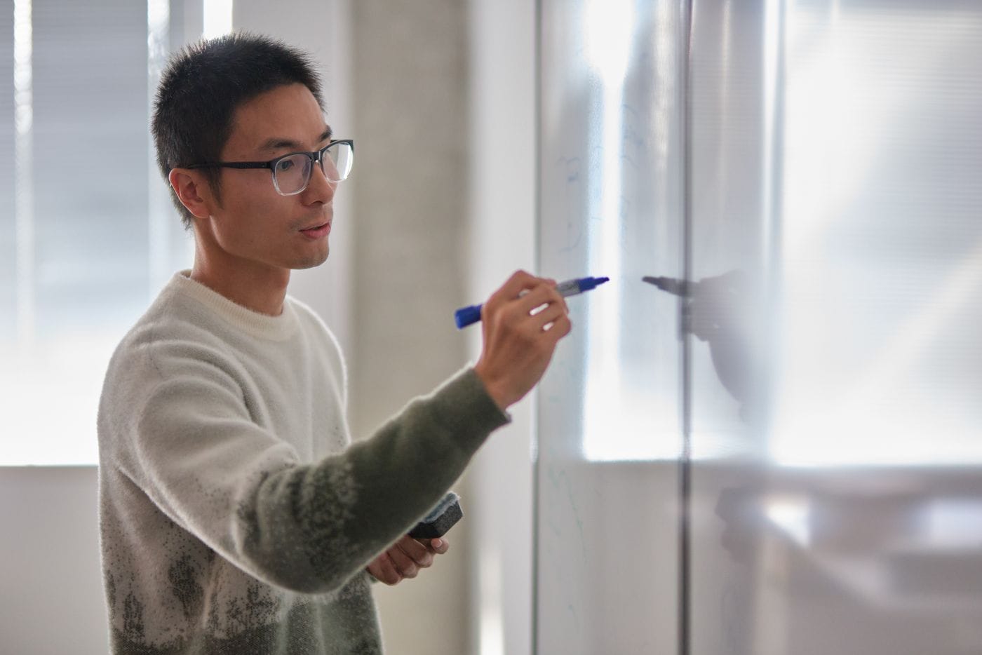 A young man with short dark hair and glasses wears a patterned beige and green sweater while writing on a whiteboard with a blue marker.