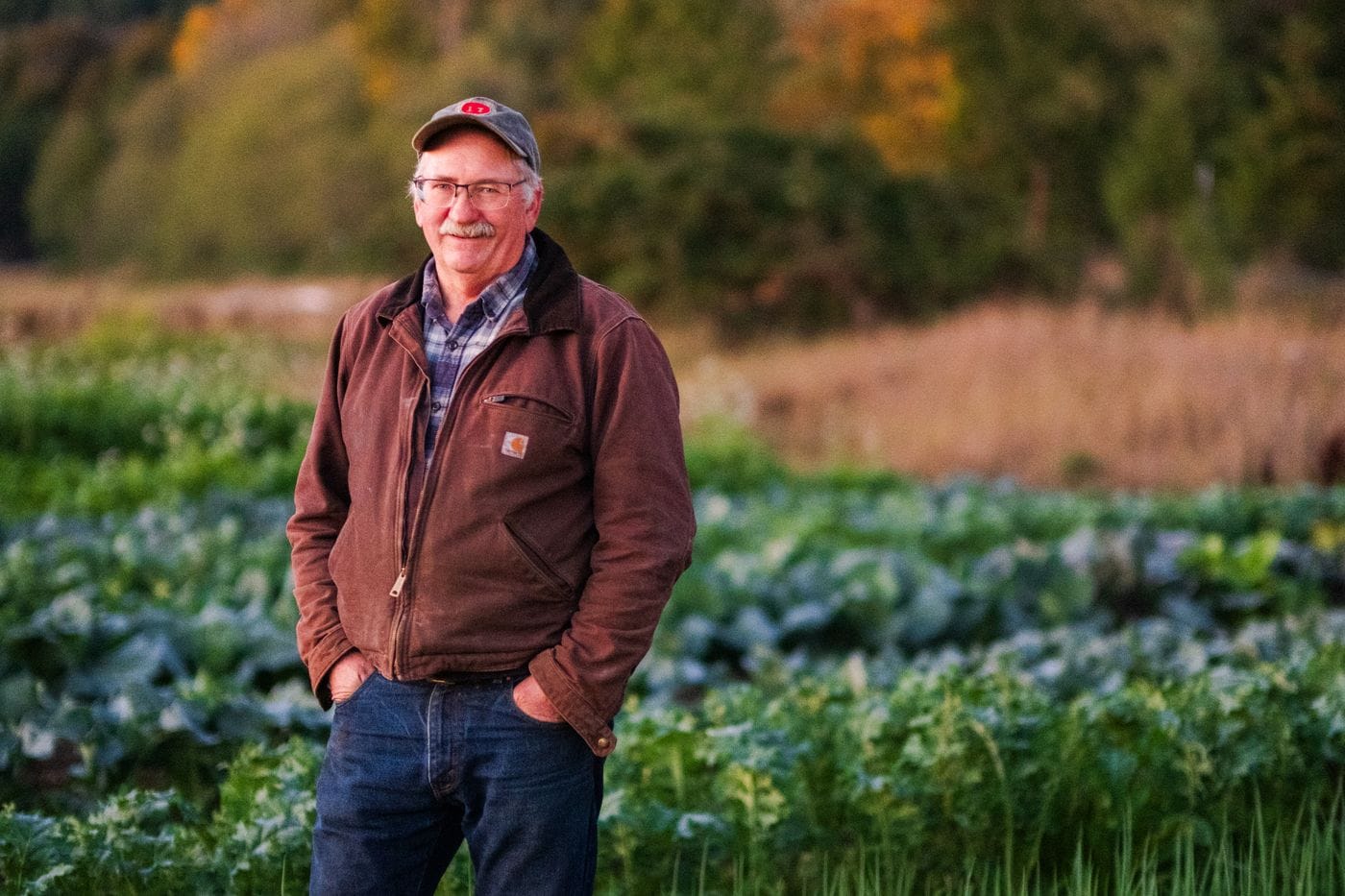 A smiling, mustachioed farmer wearing a brown Carhartt jacket, plaid shirt, jeans, and a baseball cap stands with his hands in his pockets in the middle of a vegetable field at sunset.