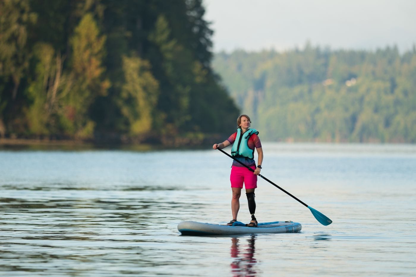 A woman wearing a life vest and pink shorts uses a paddleboard on a calm lake, with a prosthetic leg visible as she stands on the board in front of a forest shoreline.