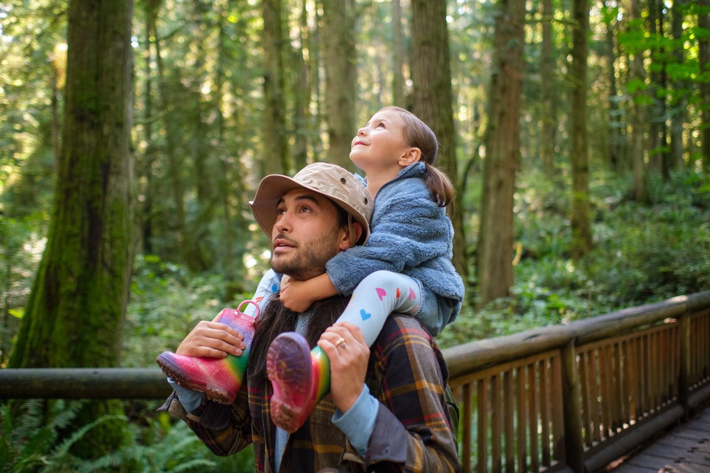 A man in a bucket hat carries a young girl on his shoulders as they both look up toward the towering trees in a sunlit forest.
