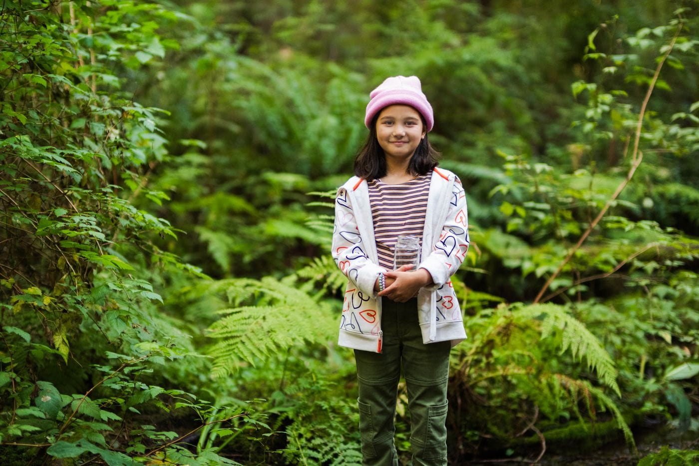 A young girl wearing a pink beanie and a heart-patterned zip-up jacket stands in a lush, green forest holding a clear glass jar.