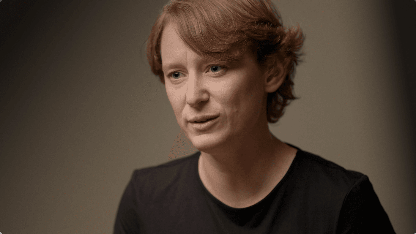 A medium close-up shot of a person with short, light brown hair and blue eyes, wearing a black shirt, looking slightly away from the camera against a plain, muted background.