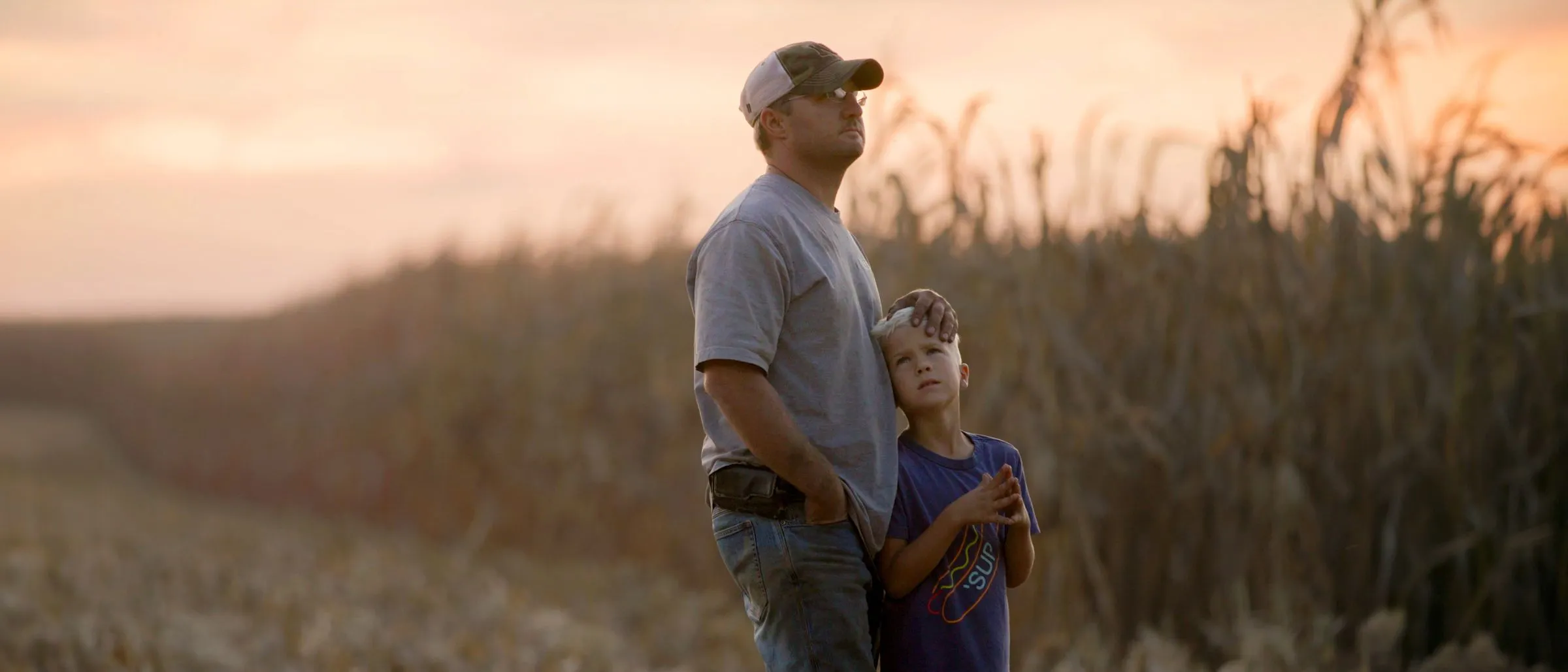 A father wearing a baseball cap and a grey t-shirt stands in a field at sunset with his hand on the head of his young son, who gazes up at him.