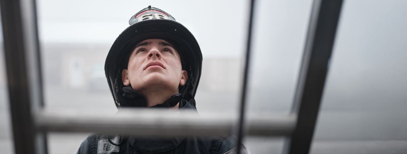 A low-angle, close-up shot of a young firefighter wearing a helmet with the number 68 as they look upward through the rungs of a ladder, set against a foggy outdoor background.