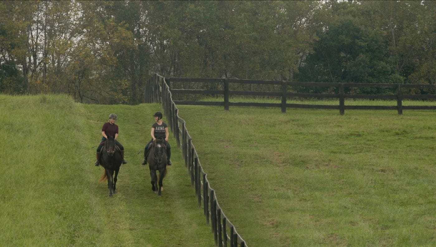 Two women wearing riding helmets ride horses side-by-side along a grassy path next to a wooden post-and-rail fence in a large, rural pasture.