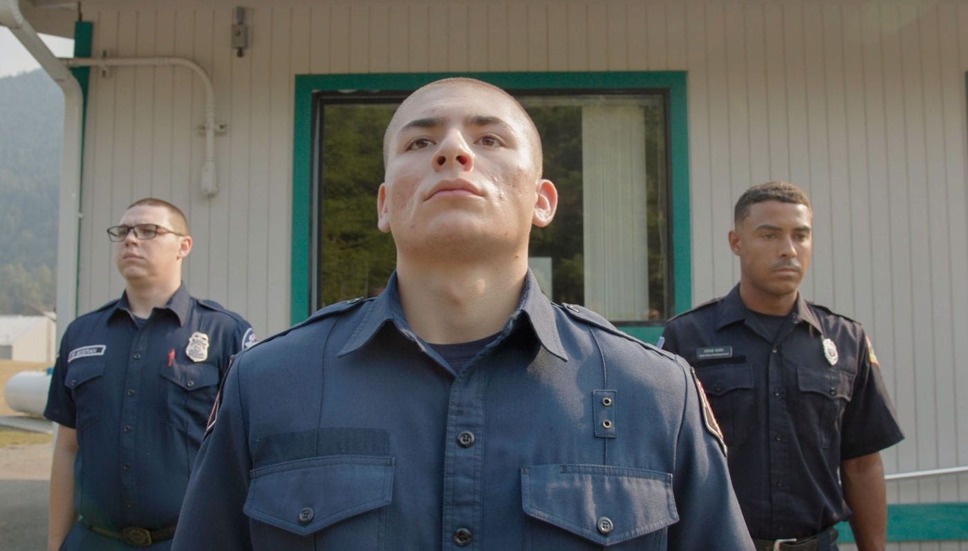 Three young men wearing dark blue police-style uniforms stand in a formal row in front of a light-colored building.