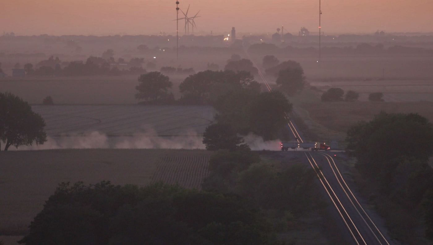 A red semi-truck pulls a trailer across a railroad crossing at dawn in a hazy, rural landscape dotted with wind turbines and agricultural fields.