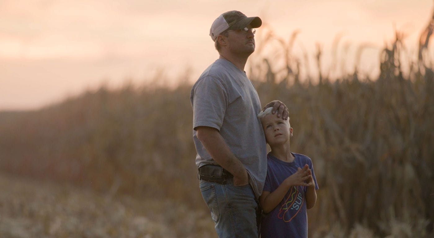 A father in a baseball cap and a young boy stand together in a field at sunset, both looking thoughtfully toward the horizon.