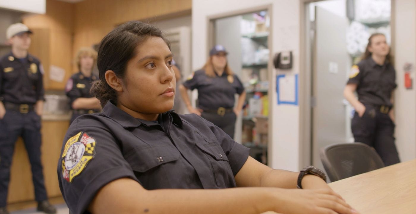 A young woman in a dark blue uniform sits at a table in a room with several other people in matching uniforms standing in the background.