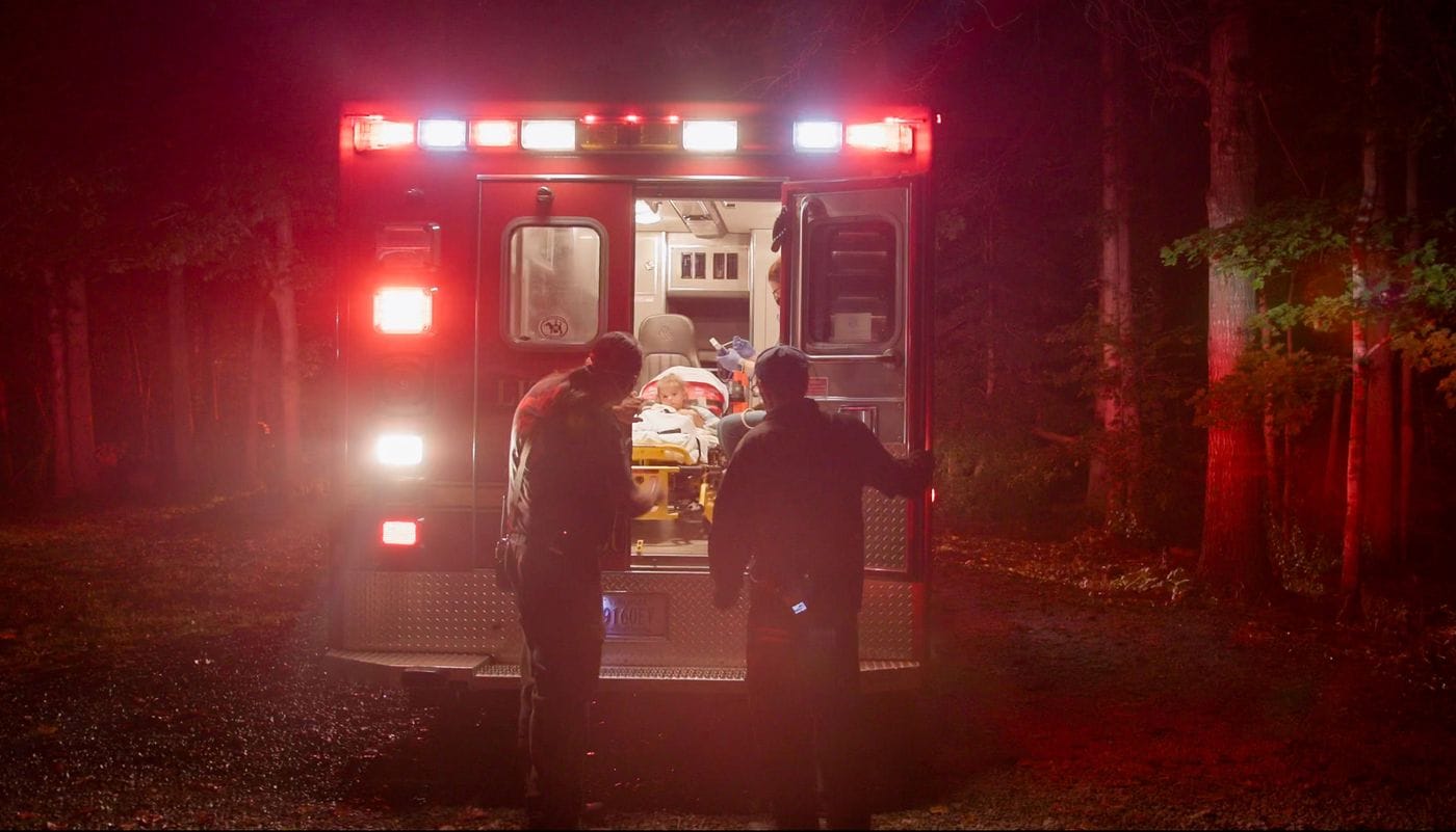 Two first responders stand at the back of an illuminated ambulance in a dark, wooded area as a young child lies on a stretcher inside the vehicle.
