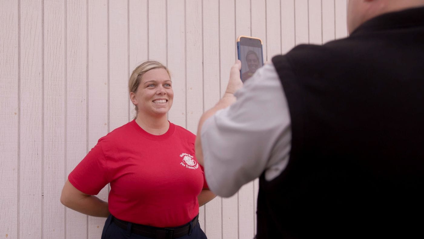A woman in a red Washington State Fire Training Academy shirt stands smiling in front of a white wall while a person in a black vest holds up a phone to take her picture.