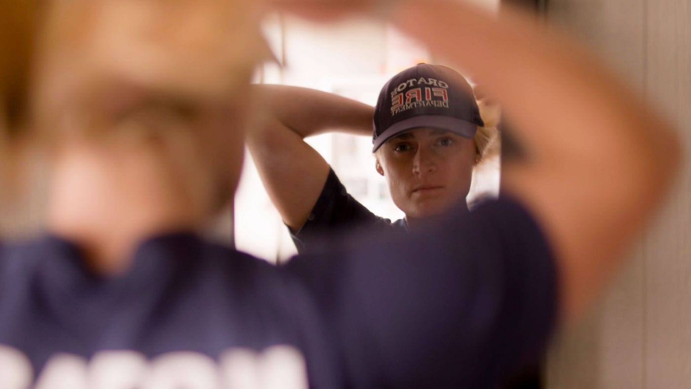 A female firefighter wearing a Gratton Fire Department baseball cap gazes at her reflection in a mirror while adjusting her hair.