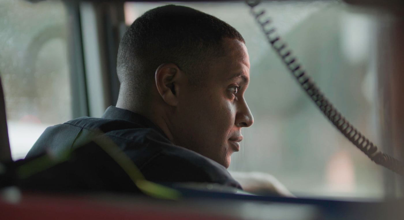 A profile view of a young Black man wearing a dark uniform, looking toward the right while sitting inside what appears to be a vehicle.