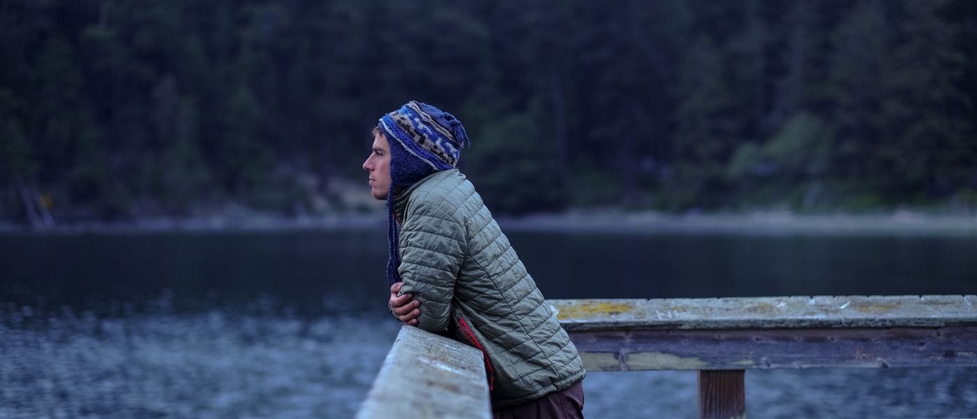 A young man in a blue patterned knit hat and olive green puffer jacket leans against a weathered wooden railing, looking out over a calm, dark lake with a forested shoreline in the background.
