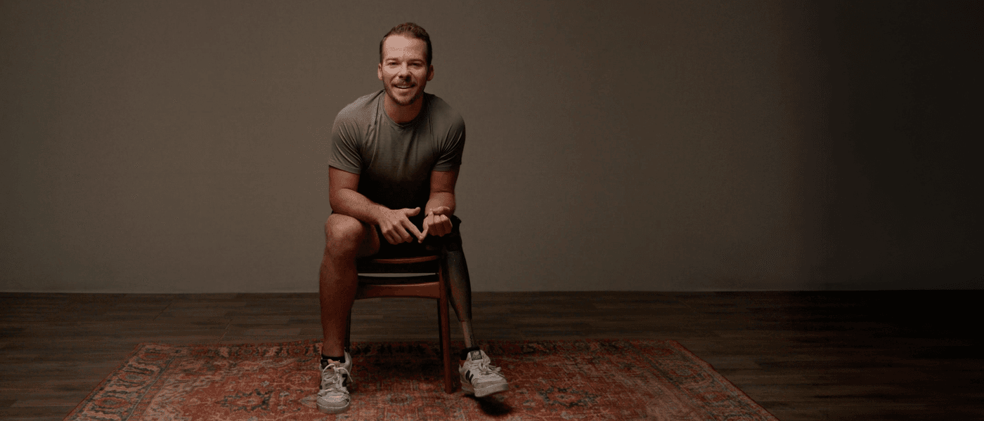 A man with a prosthetic left leg sits on a wooden chair on a patterned rug, smiling at the camera against a plain gray background.