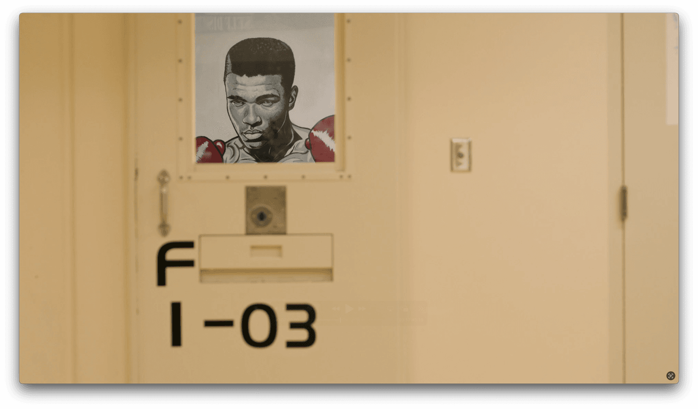 A close-up shot of a beige prison cell door labeled "F 1-03" features a portrait of boxer Muhammad Ali displayed in the small viewing window.