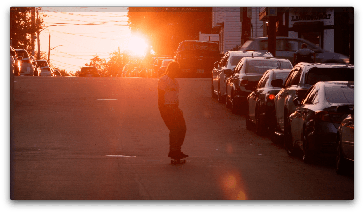 A person skateboards down the center of a city street at sunset, silhouetted against the bright golden light between rows of parked cars.