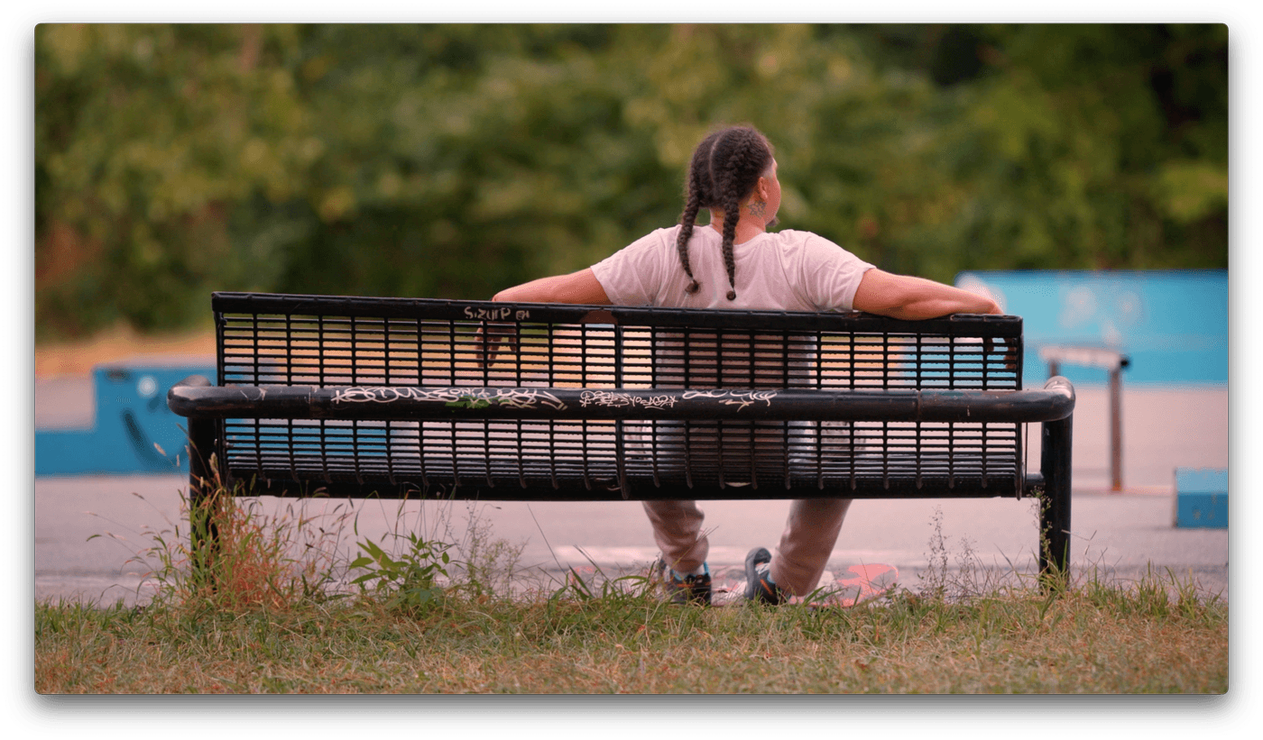 A person with braided hair sits on a black metal bench at a skatepark, viewed from behind with their arms spread across the backrest and a skateboard resting on the ground beneath them.