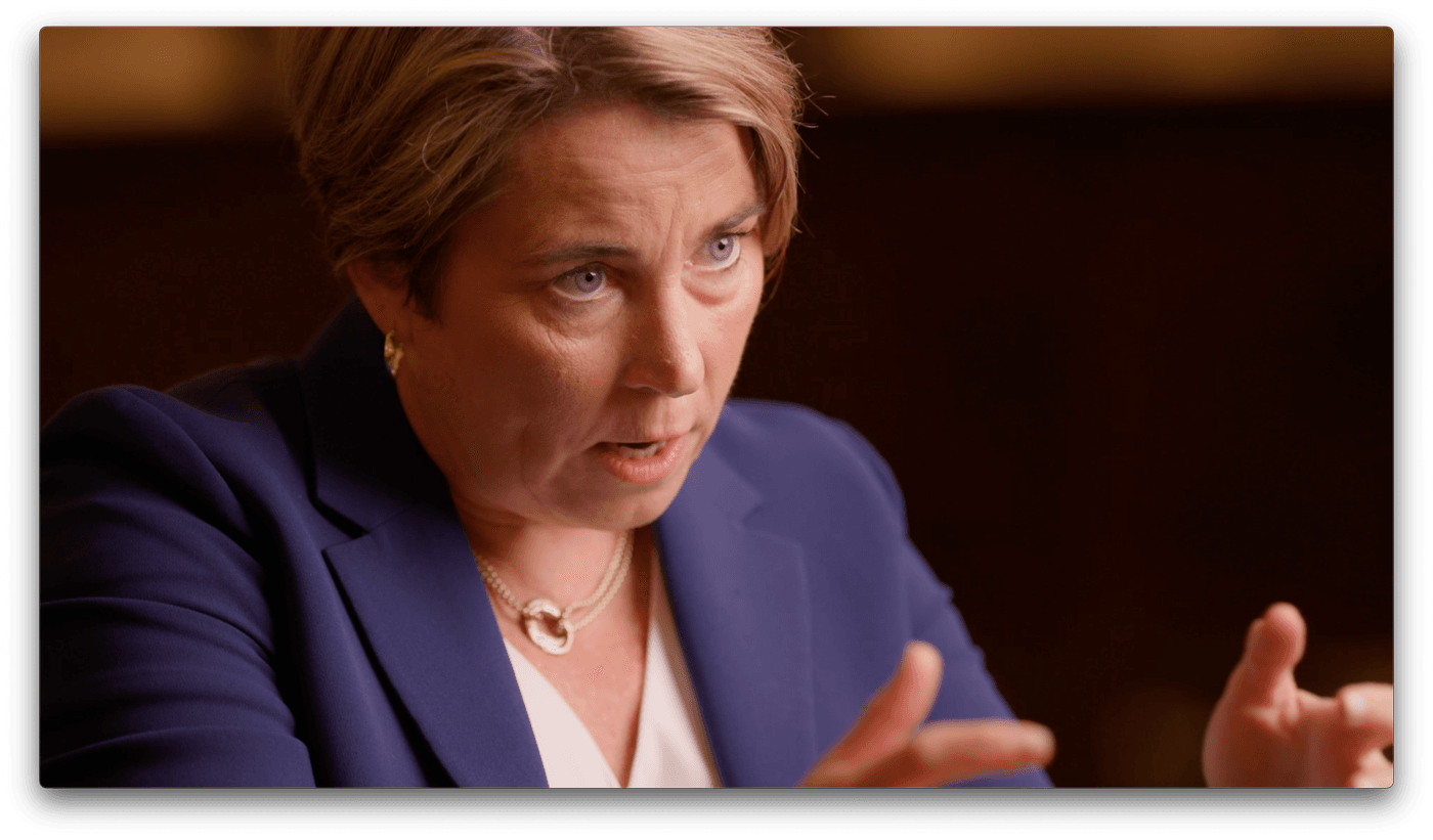 Maura Healey, wearing a blue blazer and gold necklace, gestures with her hands while speaking during a close-up interview.