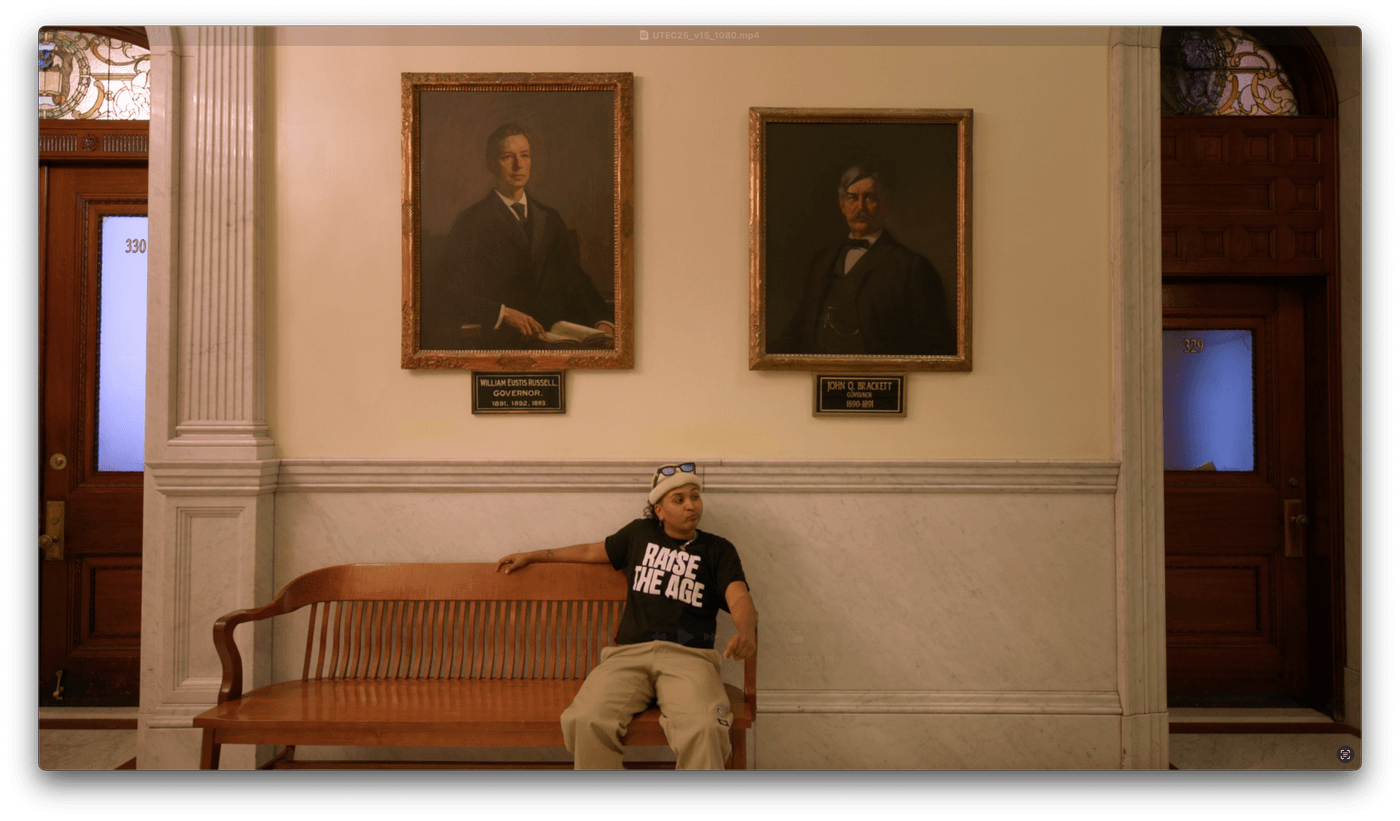 A person wearing a "RAISE THE AGE" t-shirt and a hat sits on a wooden bench in a hallway between two portraits of historical governors in a government building.