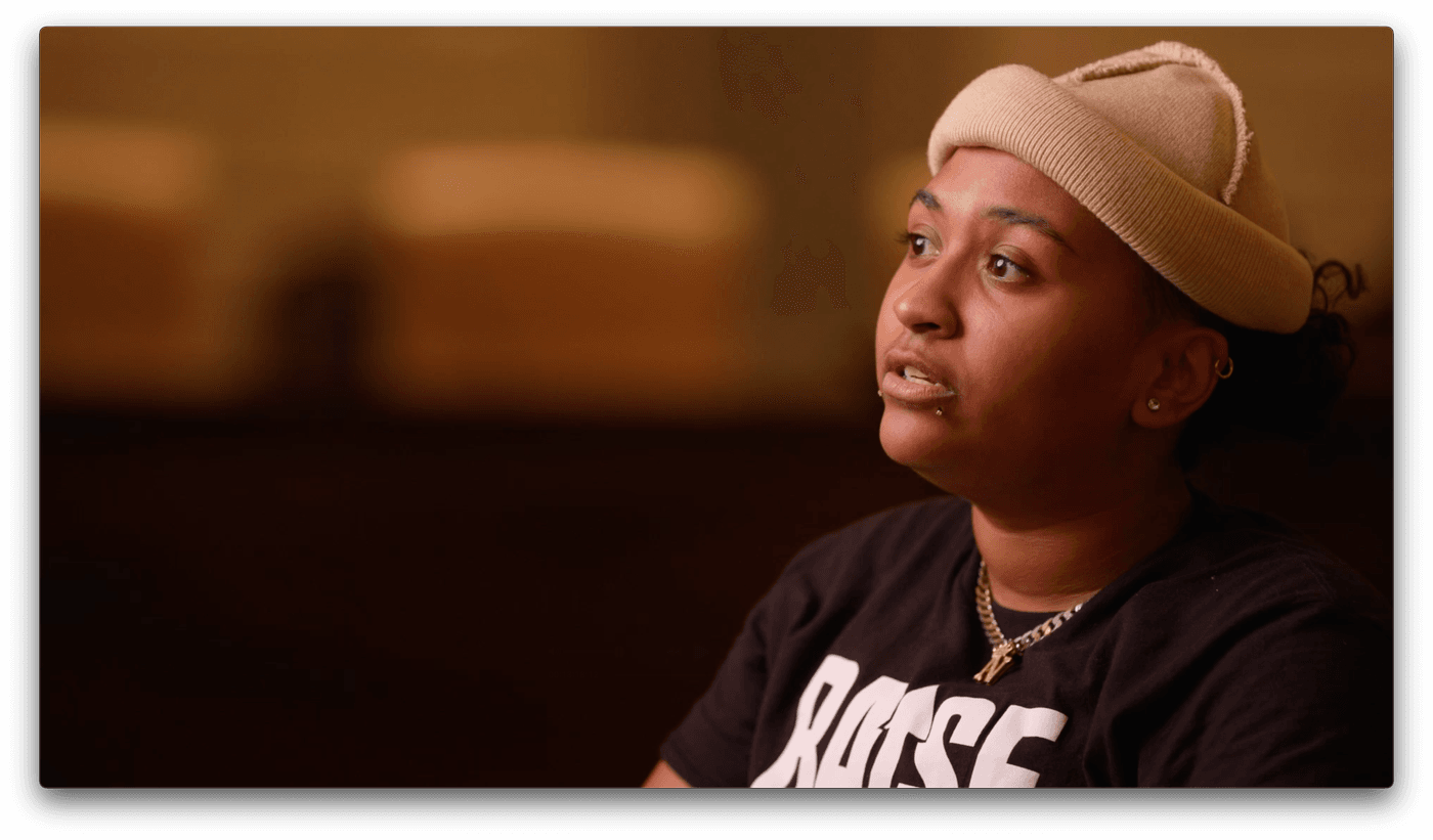 A person with short dark hair wearing a light tan beanie, black t-shirt, and silver chain necklace looks upward and to the side while speaking in a dimly lit, soft-focus setting.