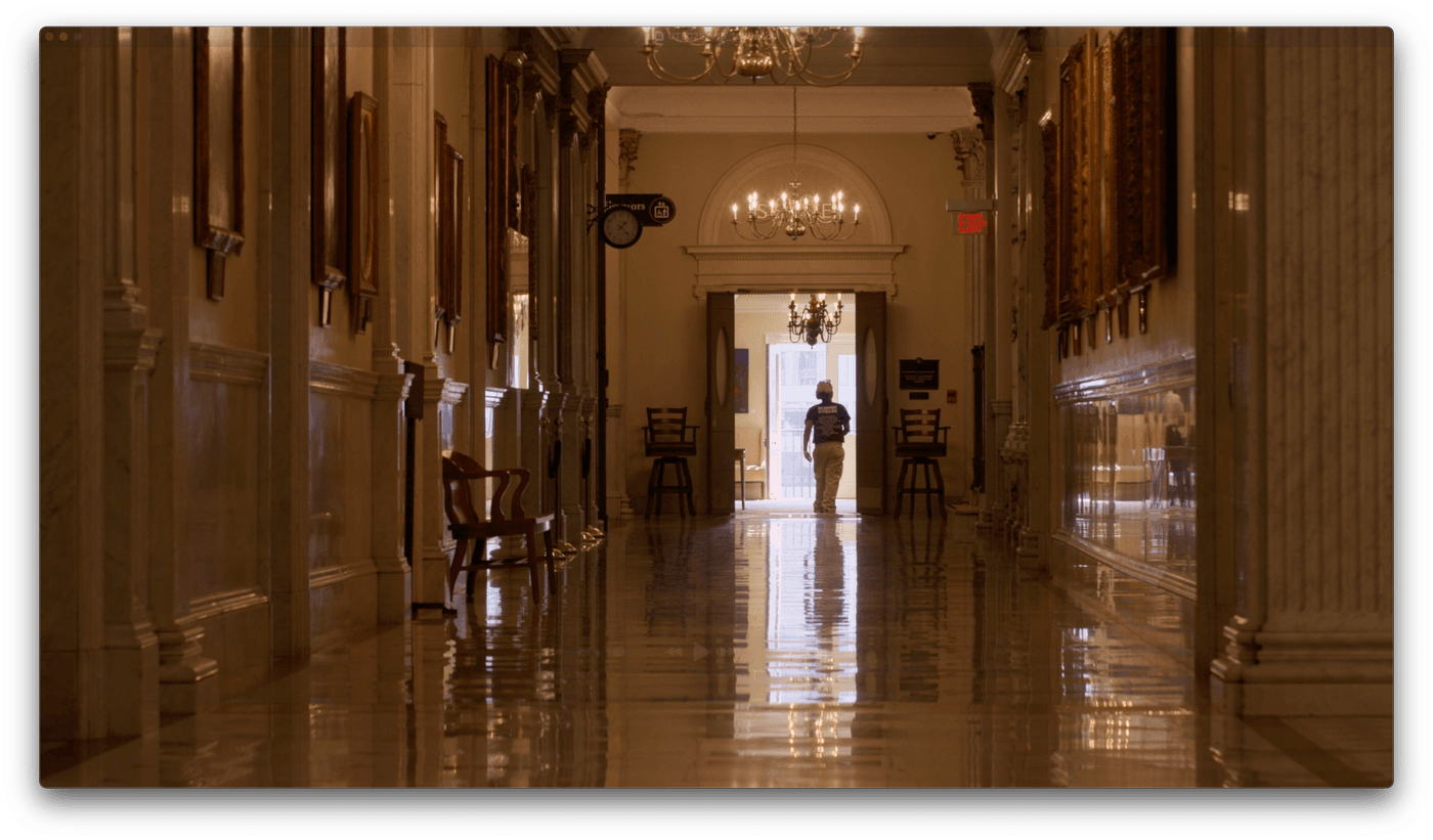 A person walks through an open doorway at the far end of a long, polished marble hallway lined with portraits and chandeliers in a grand, dimly lit building.