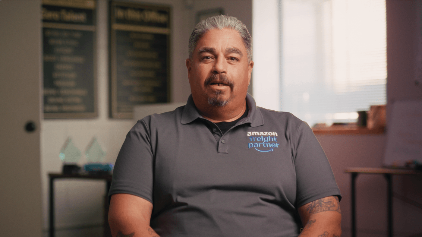 A middle-aged man with graying hair, a goatee, and tattoos on his arms wears a gray polo shirt with the Amazon Freight Partner logo while sitting in an office with plaques on the wall behind him.