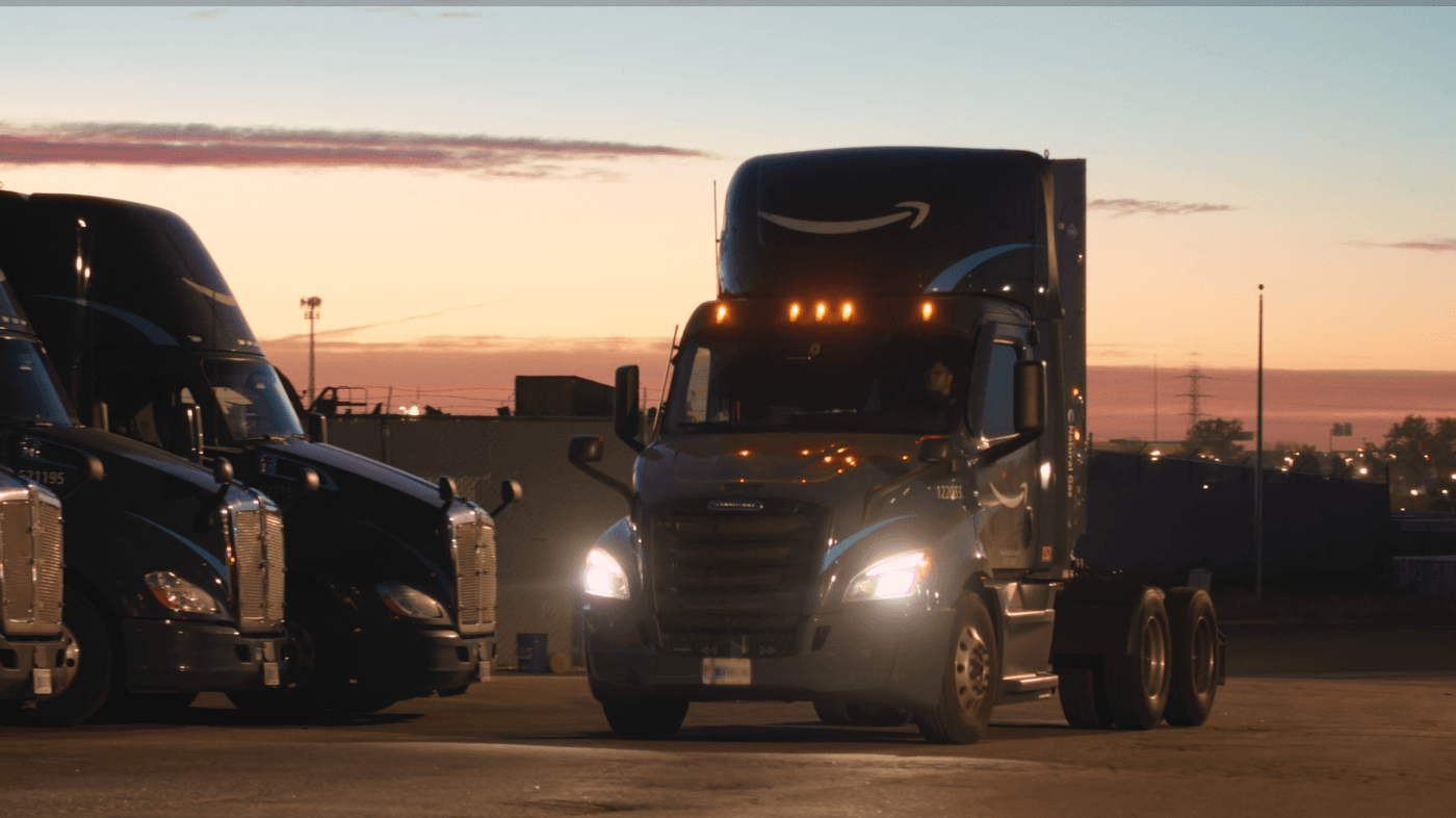 A black Amazon semi-truck with its headlights on turns in a parking lot at dawn, with two other Amazon trucks parked nearby.