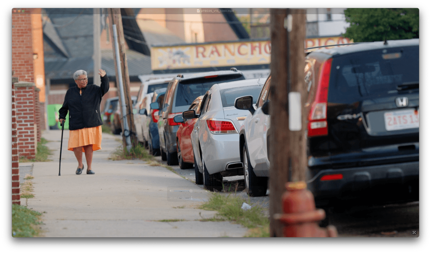 An older woman wearing a black jacket and an orange skirt waves while walking down a city sidewalk with a cane, past a row of parked cars and a restaurant sign reading "RANCHO".