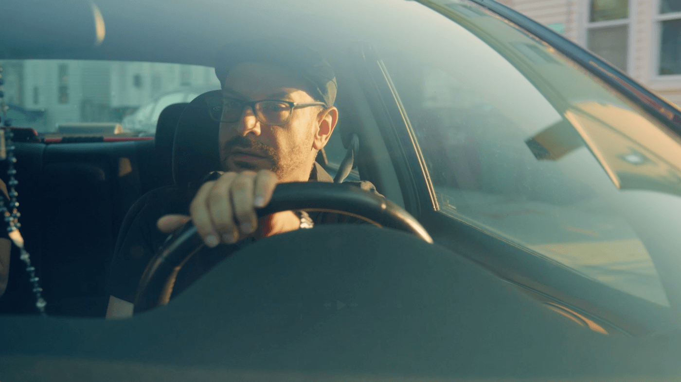 A man wearing glasses and a cap drives a car, looking toward the passenger side with a serious expression as golden hour sunlight reflects off the windshield.