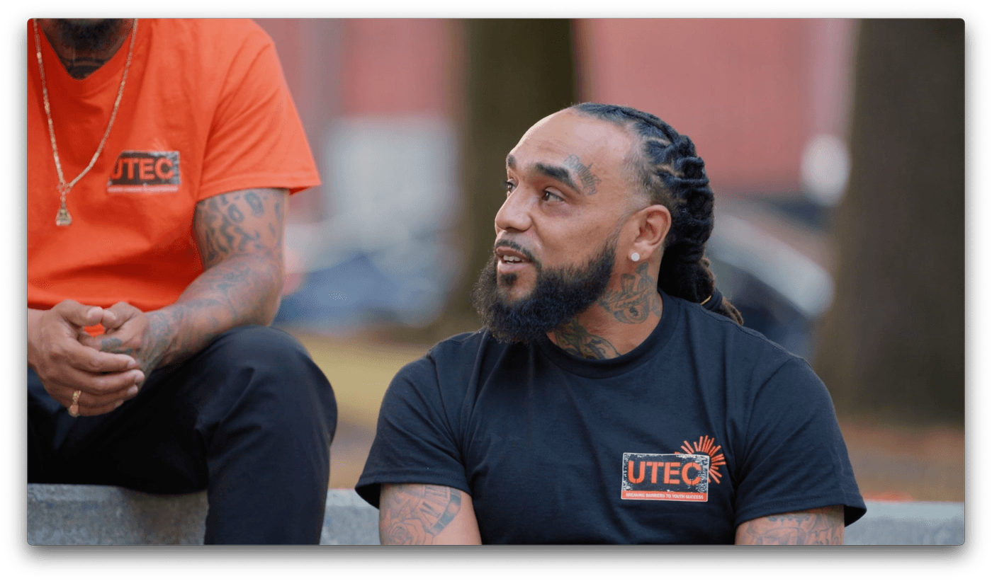 Two men with tattoos and piercings sit outdoors wearing black and orange UTEC t-shirts, with the man on the right looking toward the man on the left.
