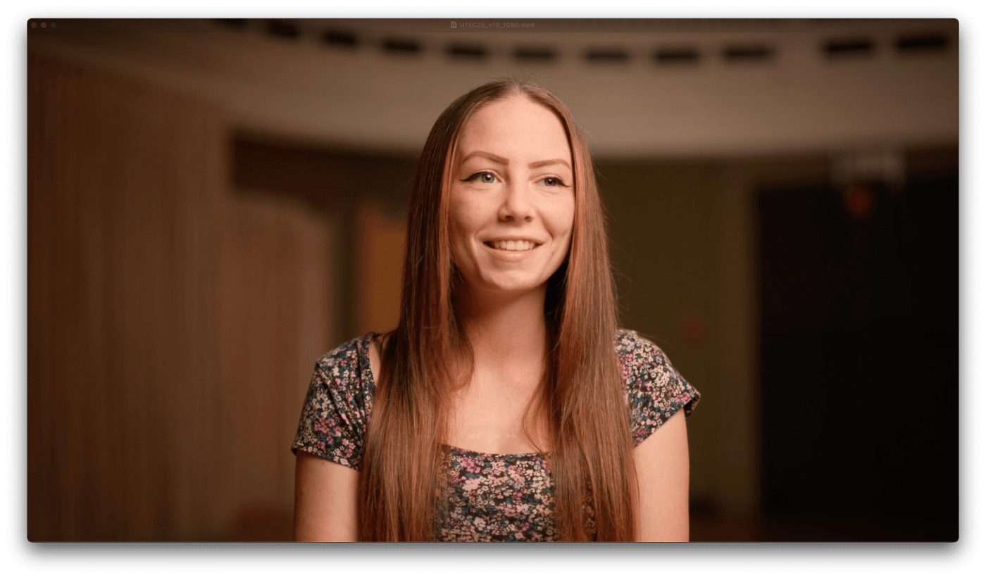A young woman with long, center-parted brown hair and eyeliner smiles while wearing a short-sleeved, multi-colored floral top in a dimly lit, soft-focus indoor setting.