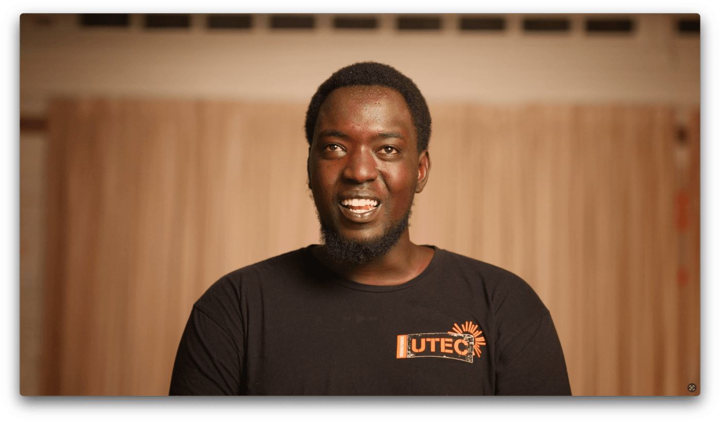 A man with short hair and a thin beard wears a black t-shirt with an orange UTEC logo as he smiles in front of beige curtains.