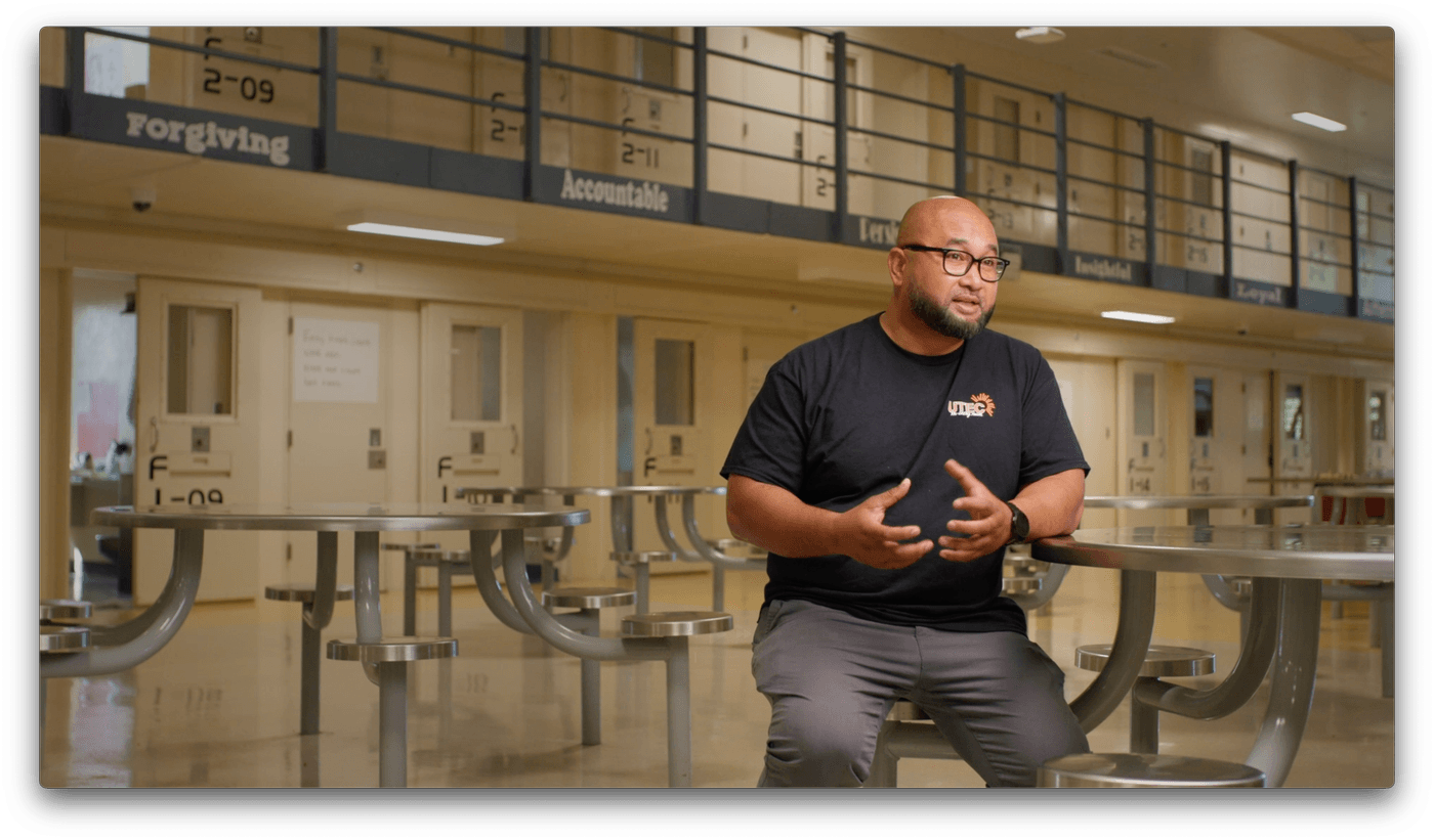 A bald man with a beard and glasses, wearing a black UTEC t-shirt, sits on a stool in a prison dayroom and gestures with his hands while speaking.