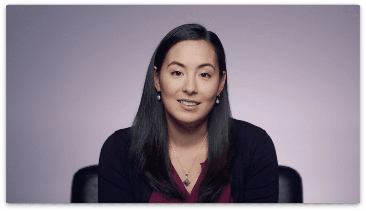 A woman with long, dark hair wearing a maroon shirt under a black cardigan smiles while looking at the camera against a soft purple background.