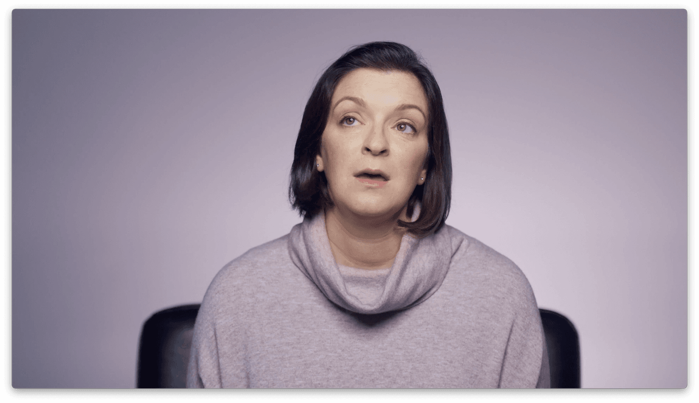 A woman with dark hair and a light pink cowl-neck sweater looks upward with a thoughtful or pensive expression against a solid light purple background.
