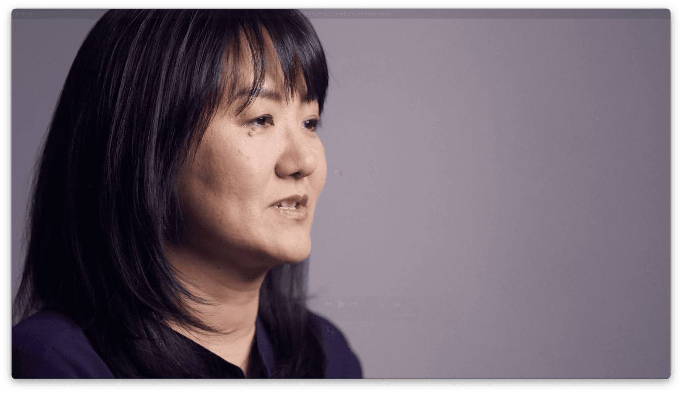 A medium shot shows a woman with dark, shoulder-length hair and bangs in profile against a plain grey background, speaking while looking off-camera.