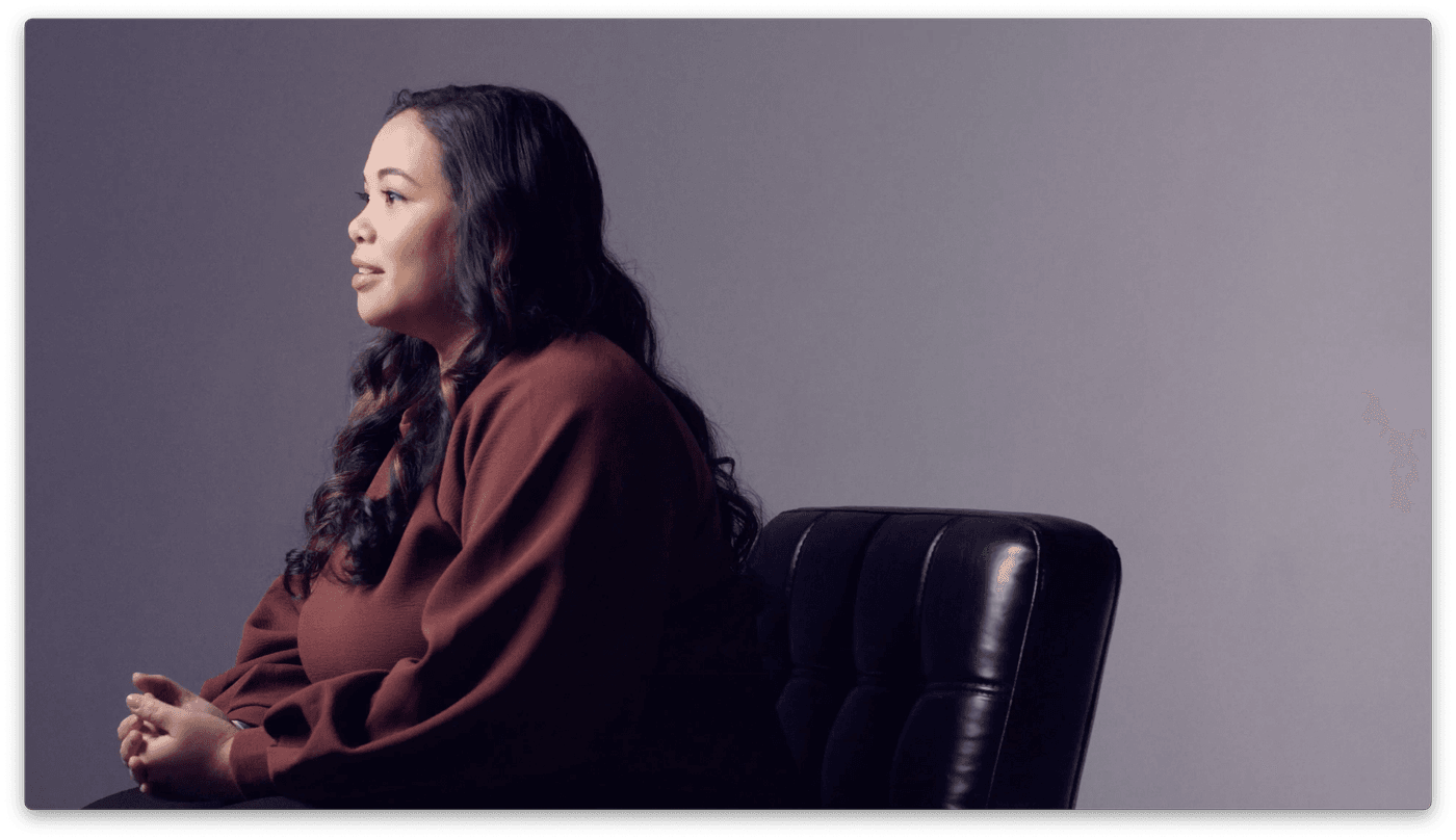 A side-profile view of a woman with long, wavy dark hair wearing a long-sleeved dark brown blouse, sitting in a black leather chair against a plain gray background.