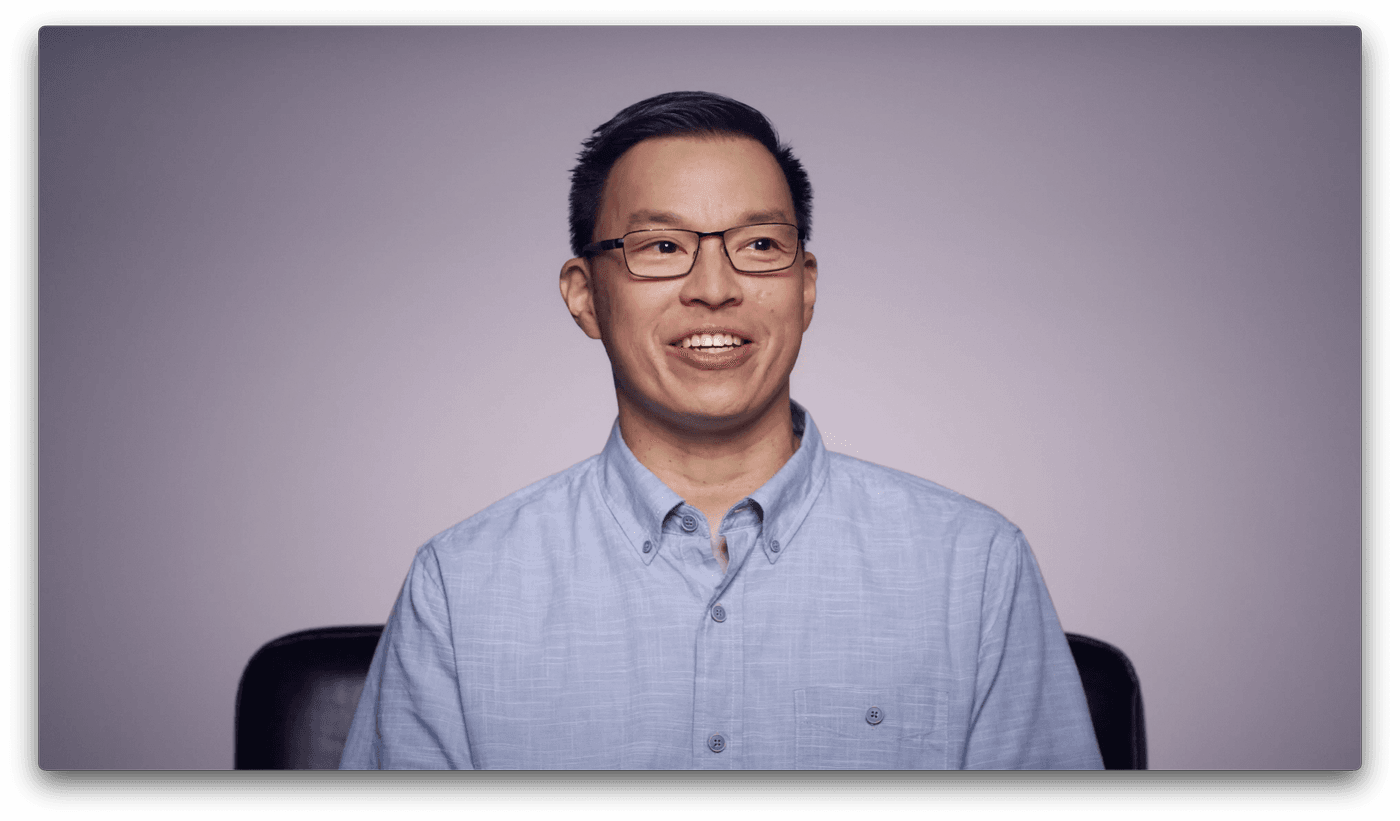 A smiling East Asian man with short dark hair and rectangular glasses wears a light blue button-down shirt against a plain, light-gray background.