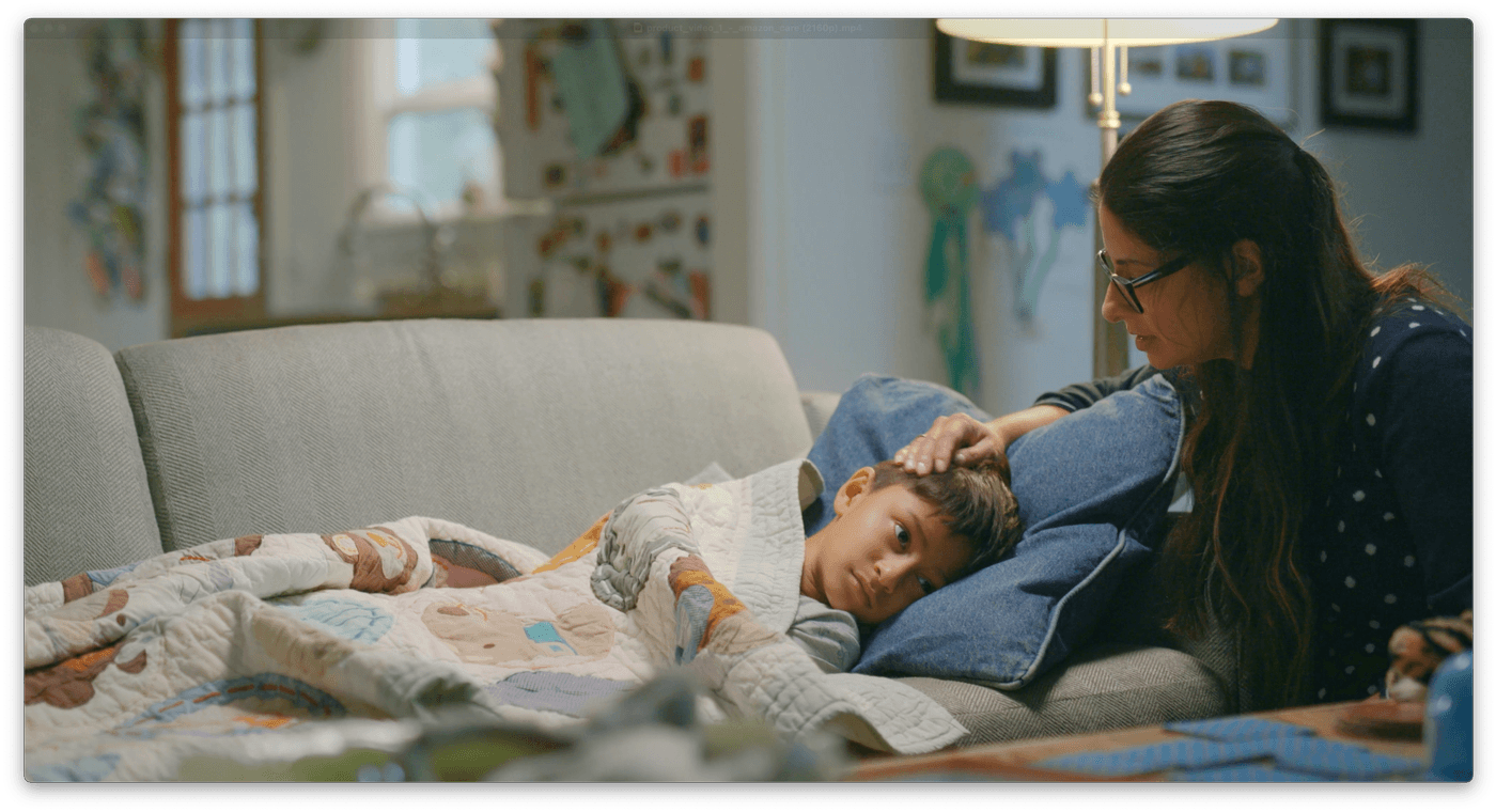 A woman with long dark hair and glasses sits on a couch, gently touching the forehead of a young boy who is lying down under a patterned quilt, appearing to comfort him.