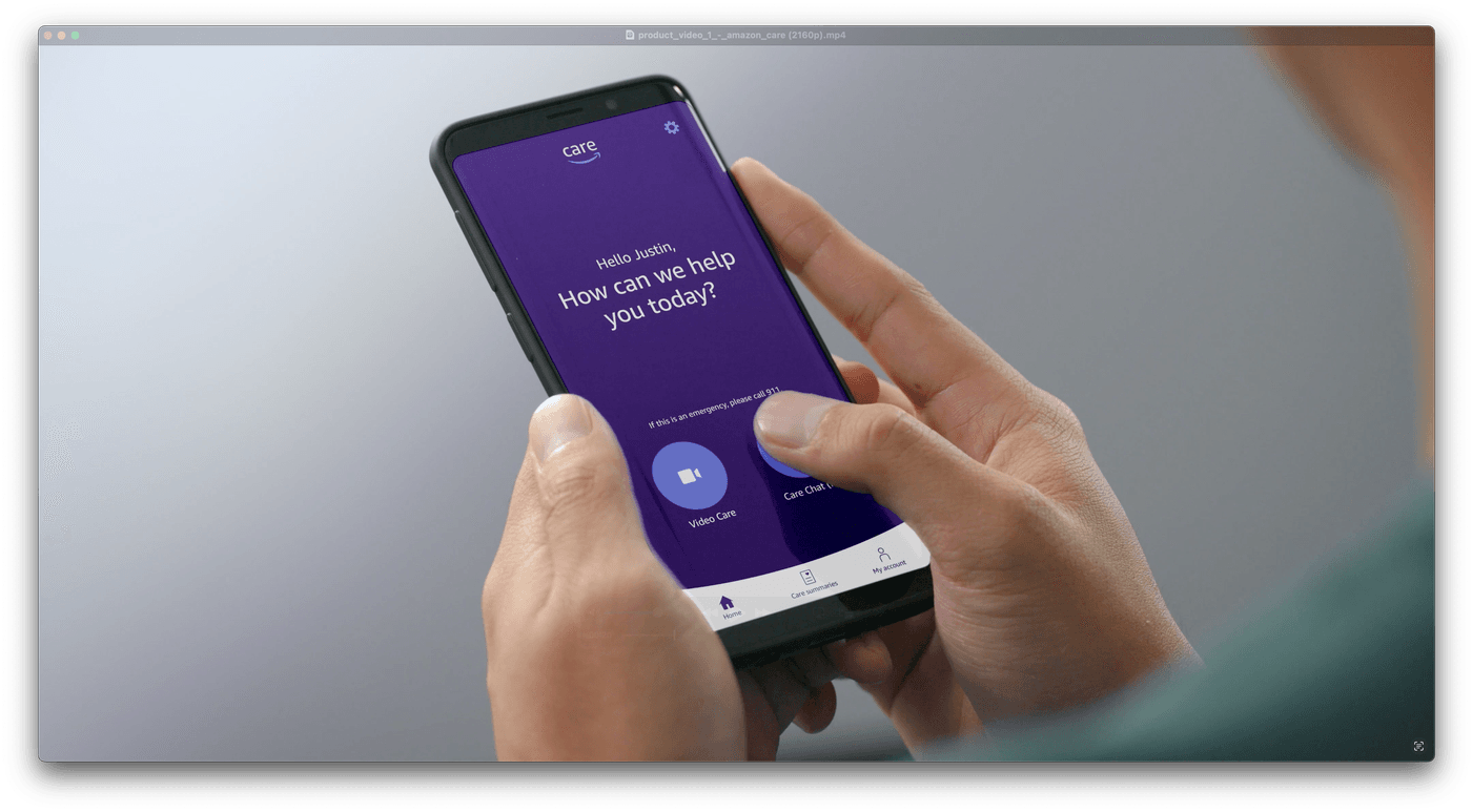 A pair of hands holds a smartphone displaying the Amazon Care mobile app interface with the greeting, "Hello Justin, How can we help you today?" and buttons for "Video Care" and "Care Chat."