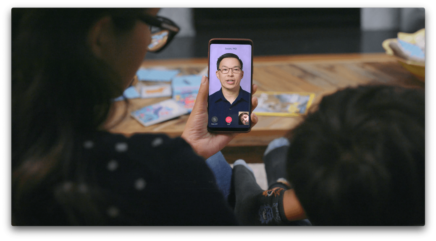 A woman wearing glasses holds a smartphone in her hand, displaying a video call with a man labeled "Jason, MD," while sitting next to a child.