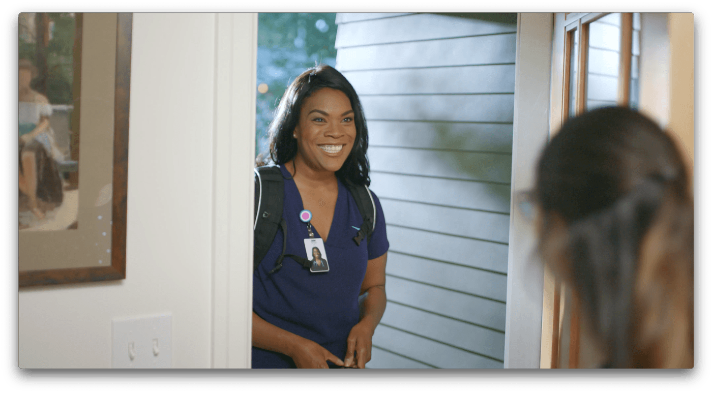 A smiling woman wearing a dark blue scrub top and a backpack with an identification badge stands at a doorway, greeting someone who is partially visible from behind.